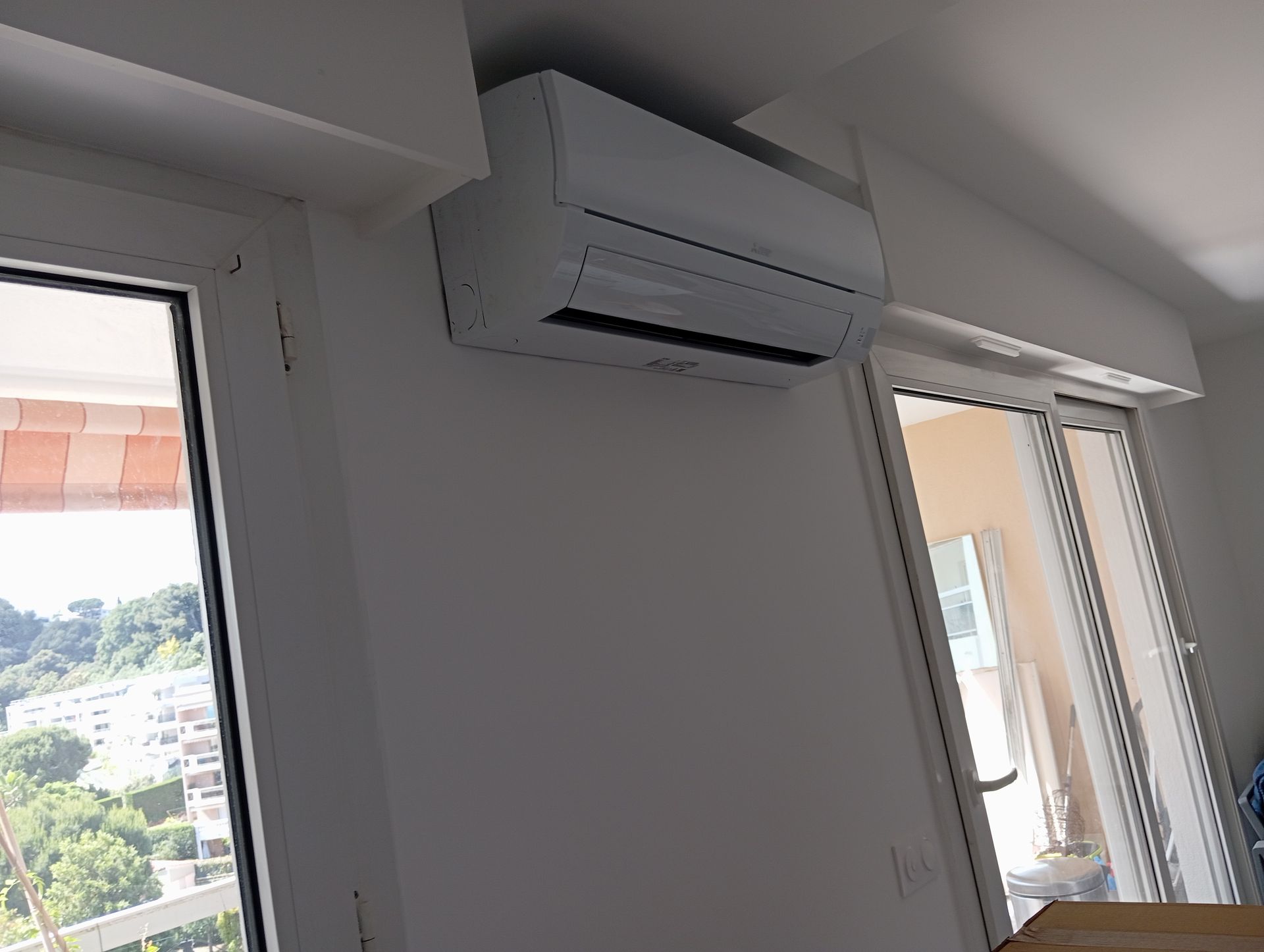 Unité de climatisation blanche montée sur un mur blanc entre deux fenêtres, donnant sur un paysage urbain.