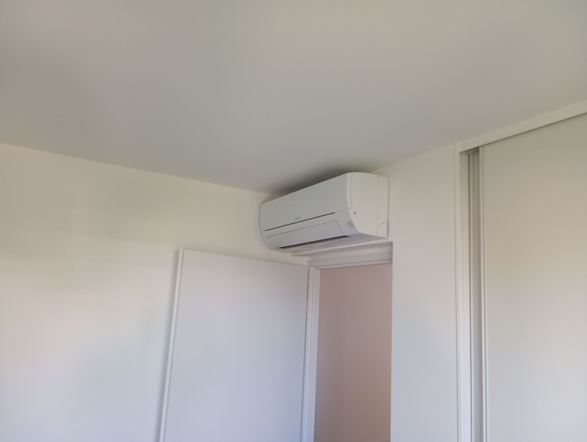 Climatiseur blanc monté sur un mur blanc au-dessus d'une porte ouverte, à l'intérieur d'une pièce.