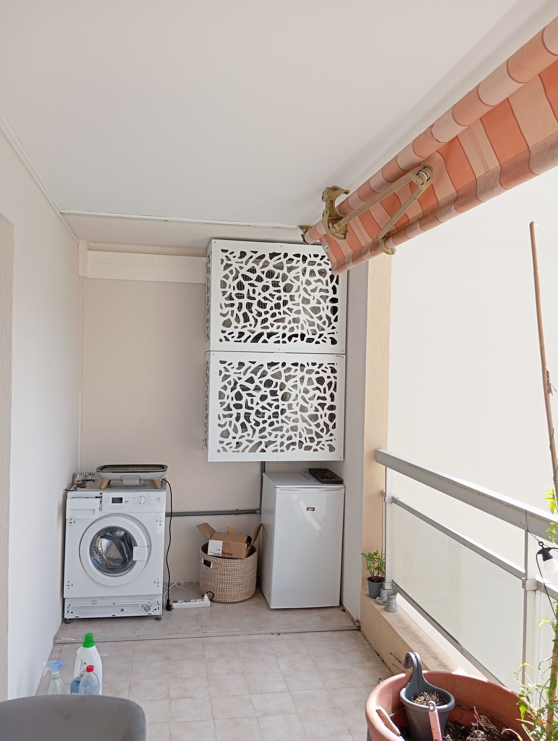 Espace buanderie sur un balcon : laveuse, sécheuse, rangement et une plante en pot sous un auvent rayé.