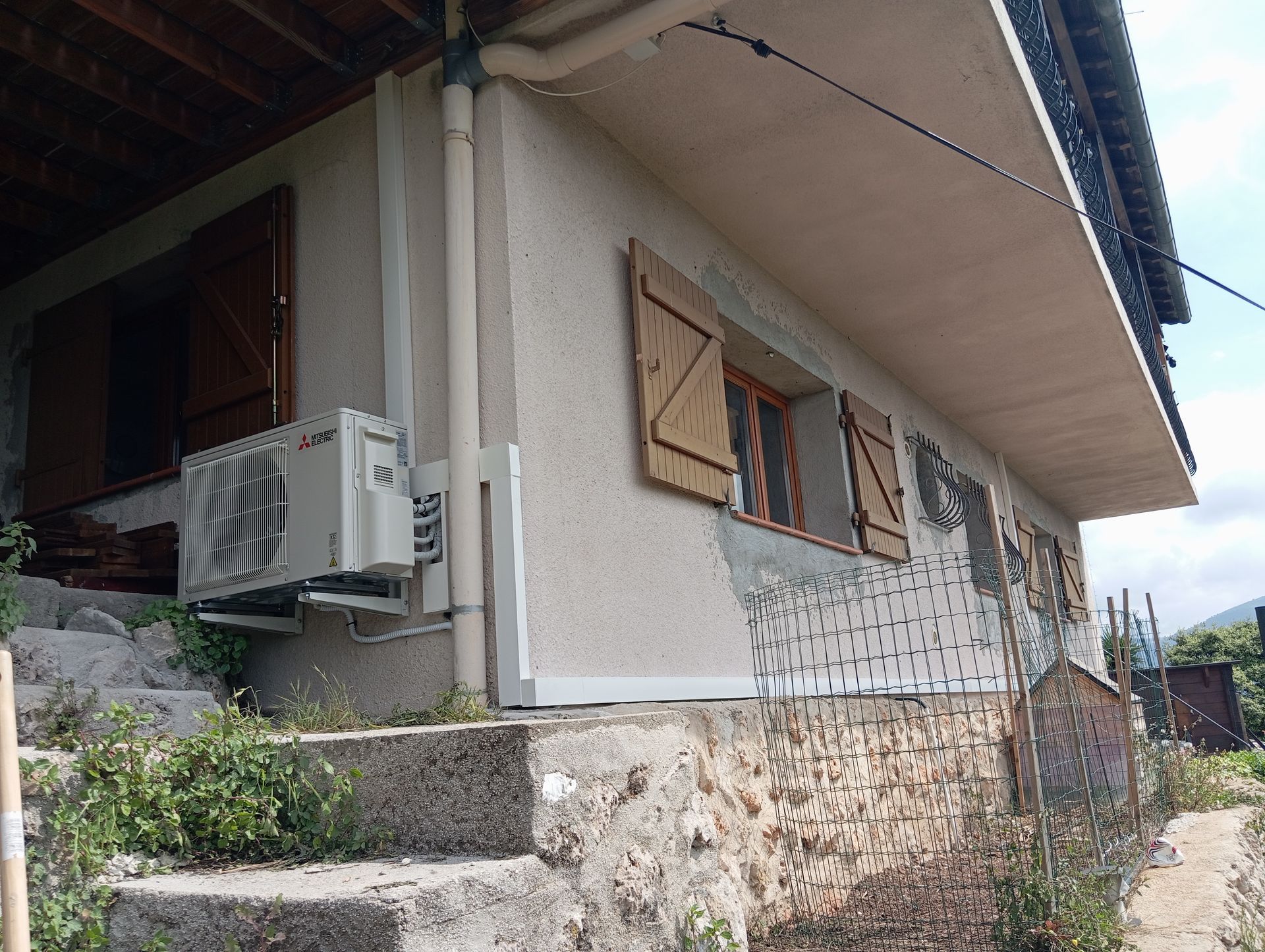 Vue latérale d'un bâtiment de couleur claire avec fondation en pierre, volets marron et unité de climatisation.