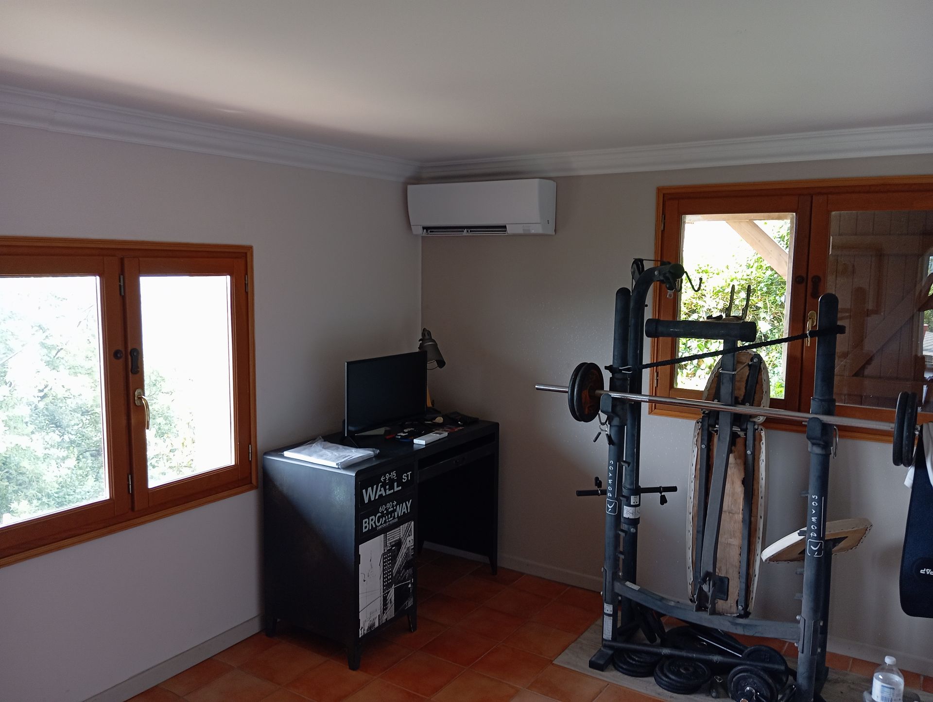 Salle de sport à domicile avec support de musculation, bureau avec ordinateur et climatisation. Deux fenêtres offrent une lumière naturelle.