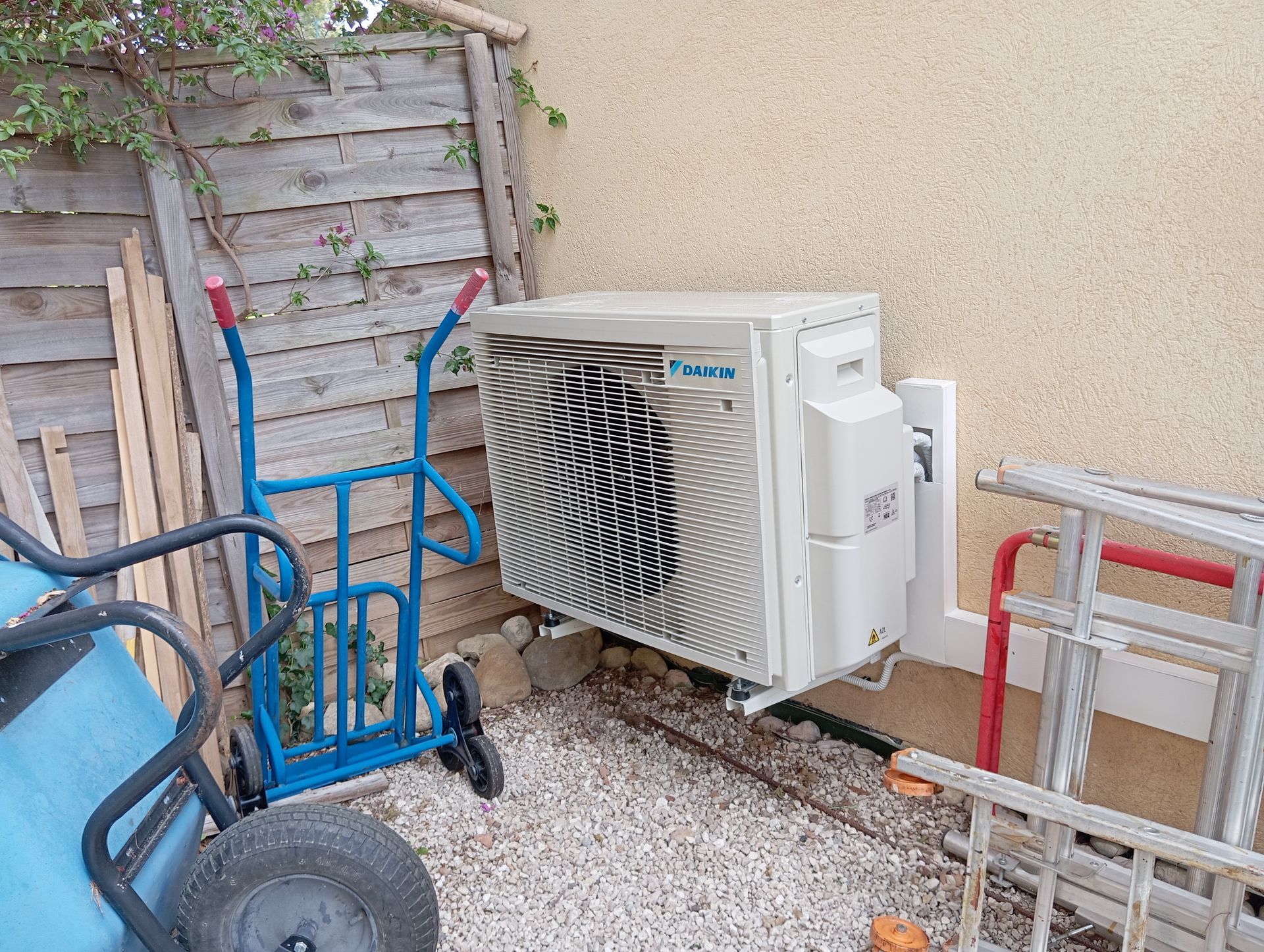 Climatiseur extérieur Daikin monté sur un mur beige, à côté d'une clôture en bois, d'un diable et d'une échelle.