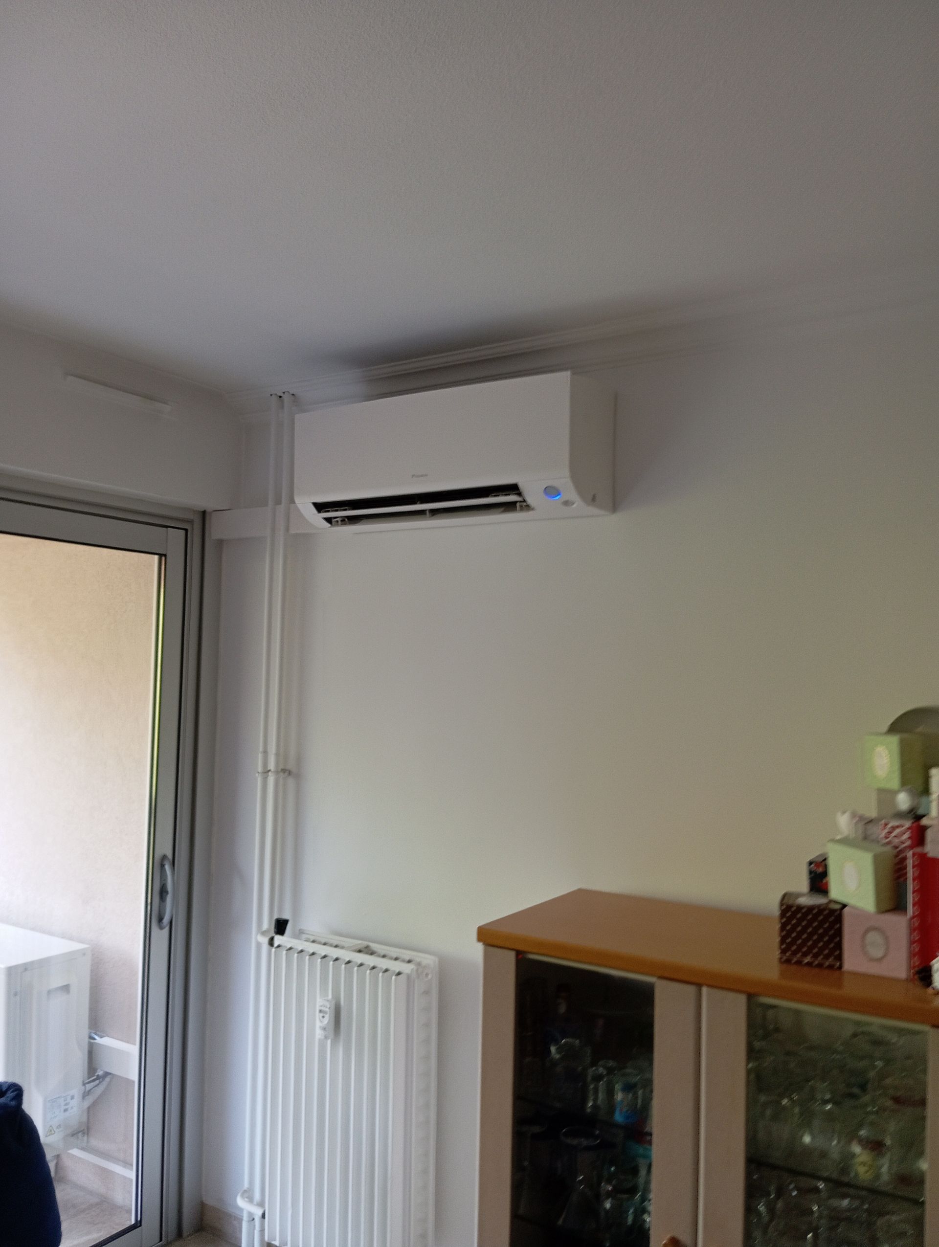 Unité de climatisation montée sur un mur blanc au-dessus d'un radiateur et d'une armoire, à côté d'une porte de balcon.