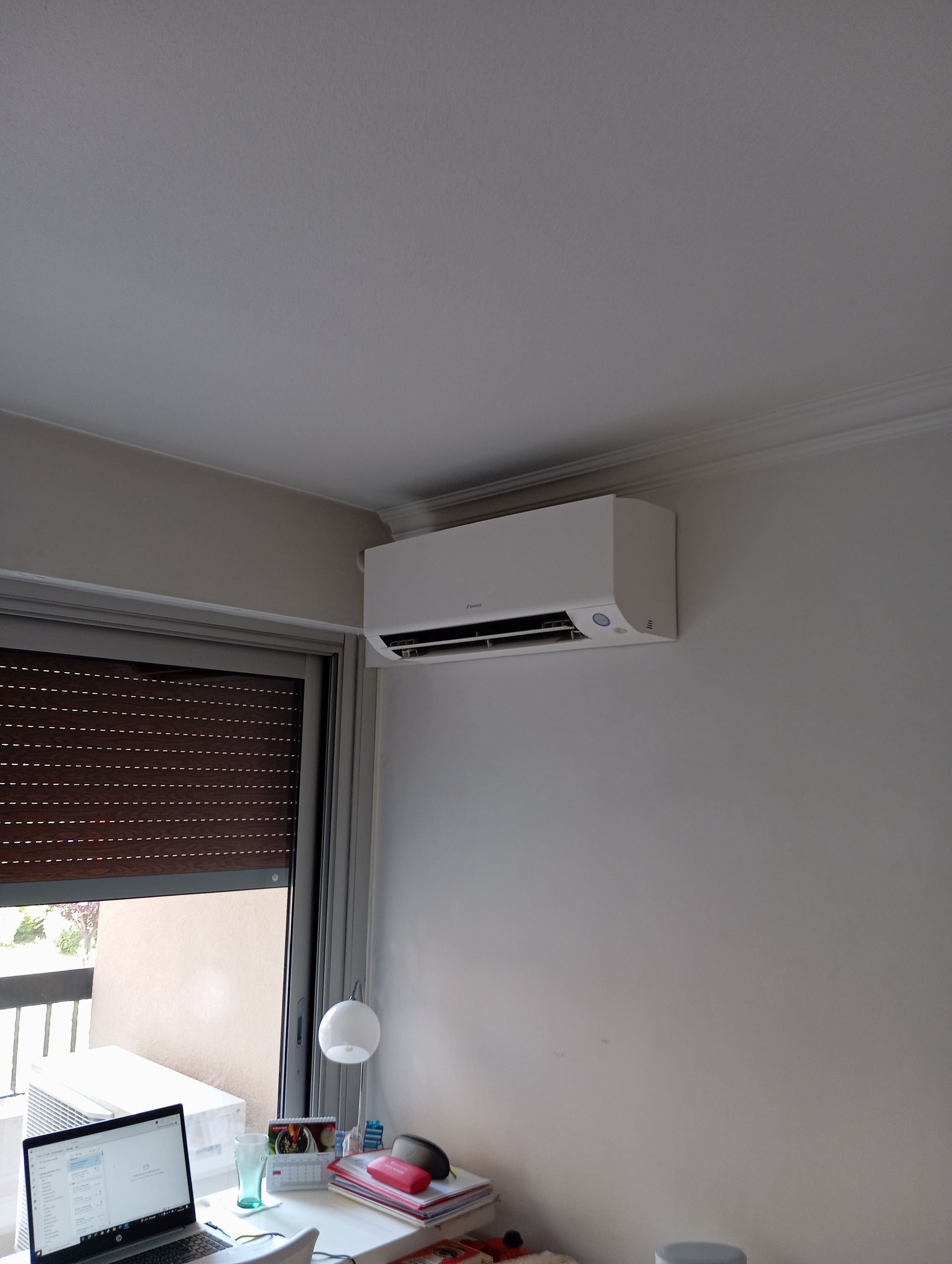 Climatiseur blanc sur un mur beige au-dessus d'un bureau avec un ordinateur portable et une fenêtre avec un store marron fermé.