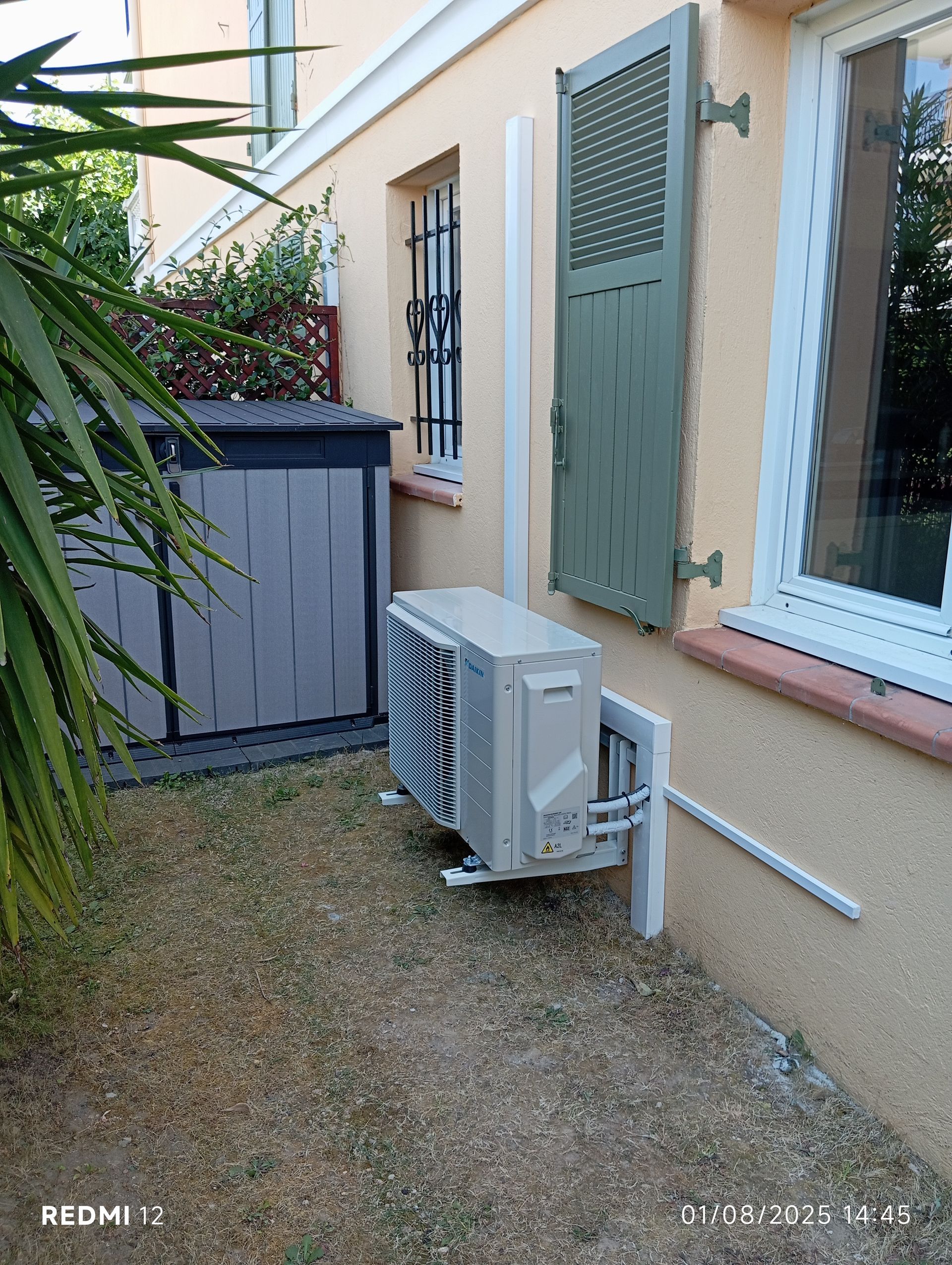 Unité de climatisation sur gravier à côté d'un bâtiment avec volets et petites fenêtres ; un conteneur de stockage se trouve à proximité.