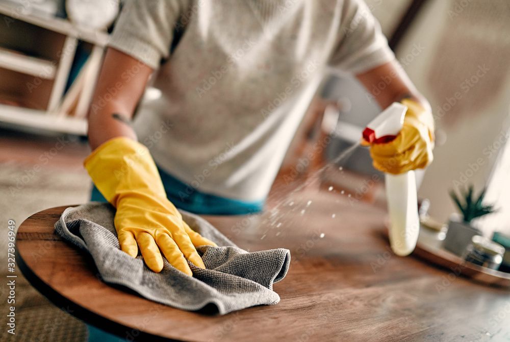 Une personne portant des gants jaunes nettoie une table en bois avec un chiffon et un vaporisateur.