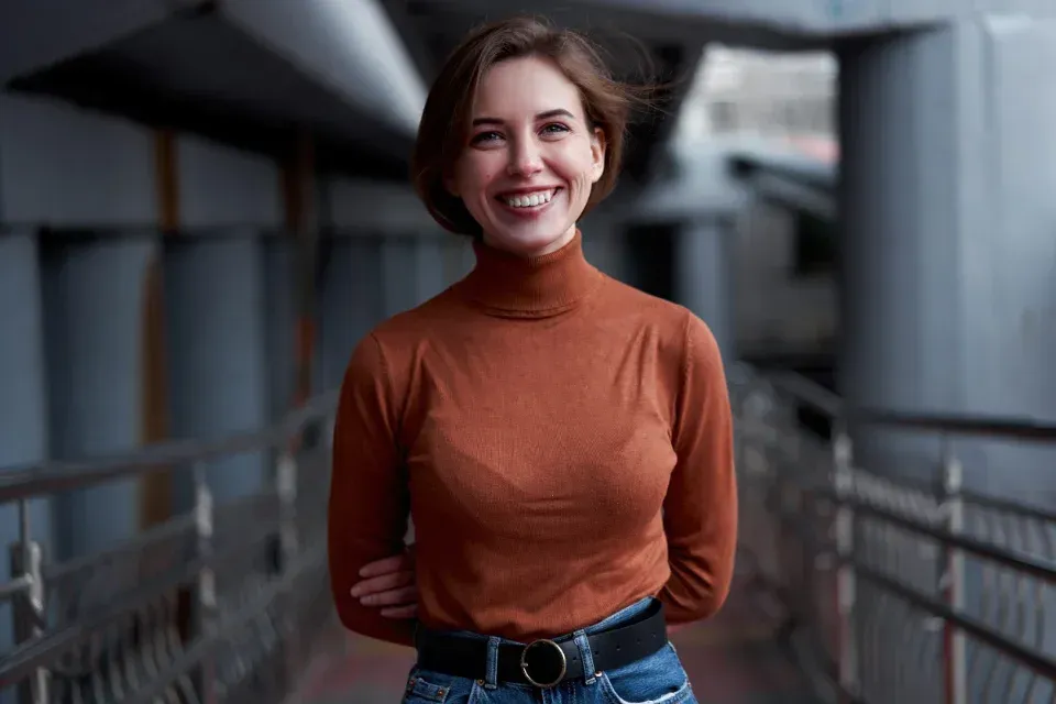 Persona sonriente con jersey de cuello alto naranja y vaqueros de pie sobre la barandilla de un puente.