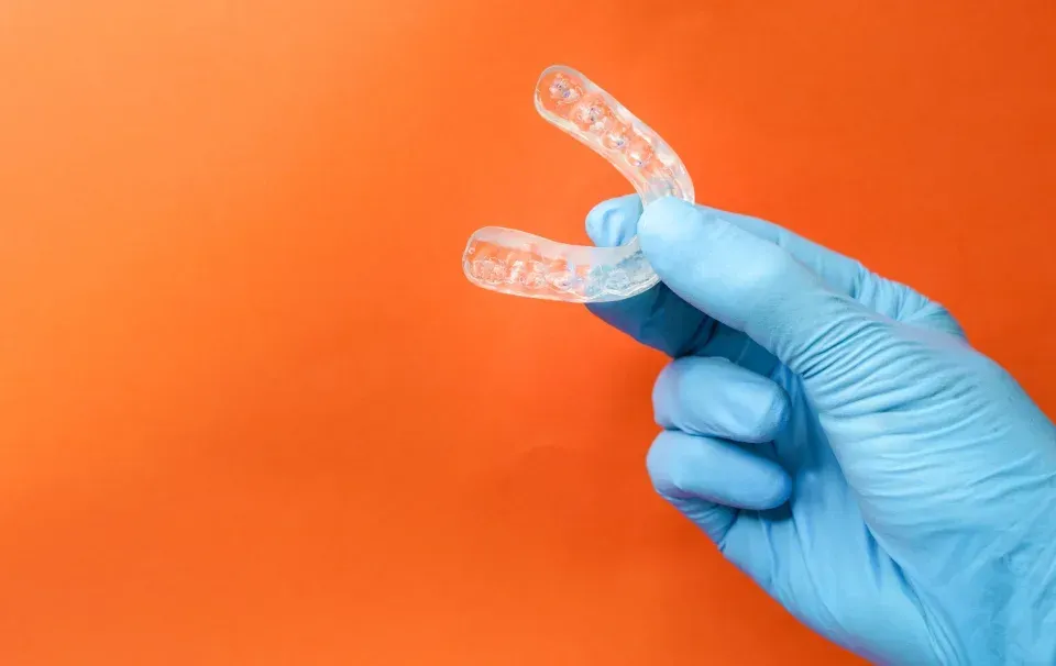 Mano enguantada de azul sosteniendo alineadores dentales transparentes sobre un fondo naranja.