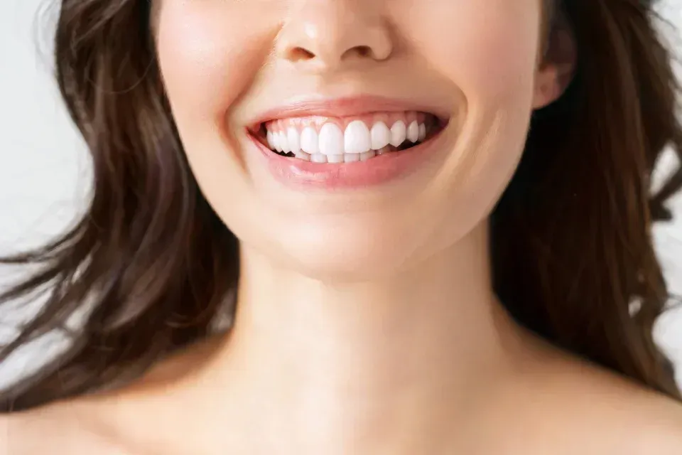 Primer plano de una persona sonriente con dientes blancos y cabello oscuro sobre un fondo claro.