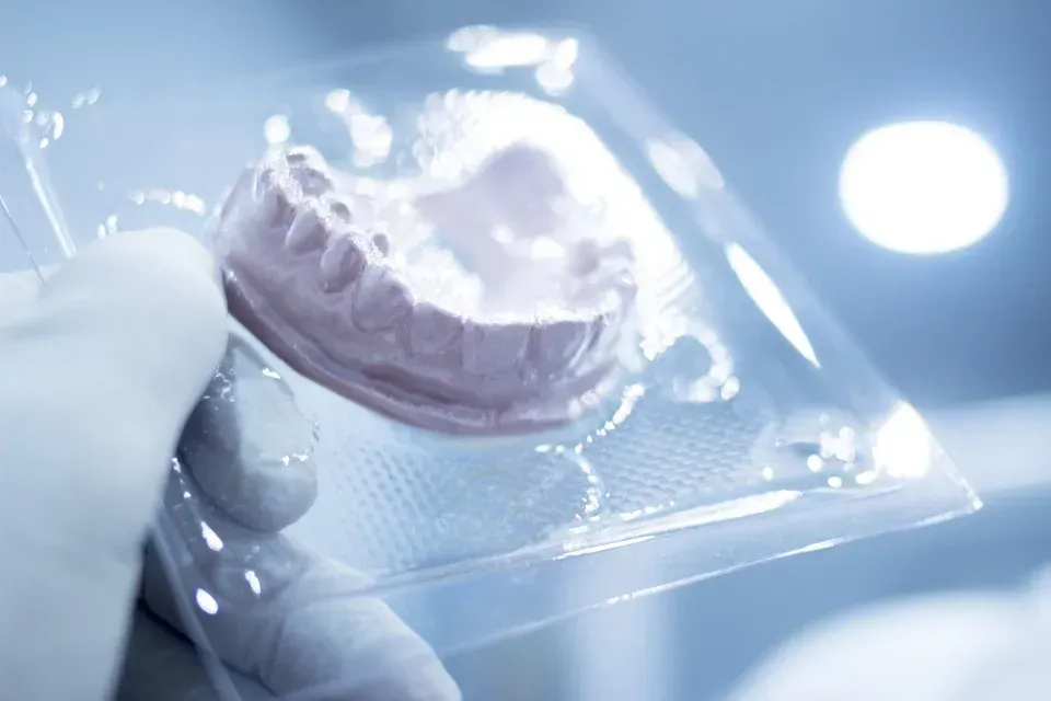 Mano enguantada sosteniendo un paquete de plástico transparente con un molde dental bajo una luz brillante.