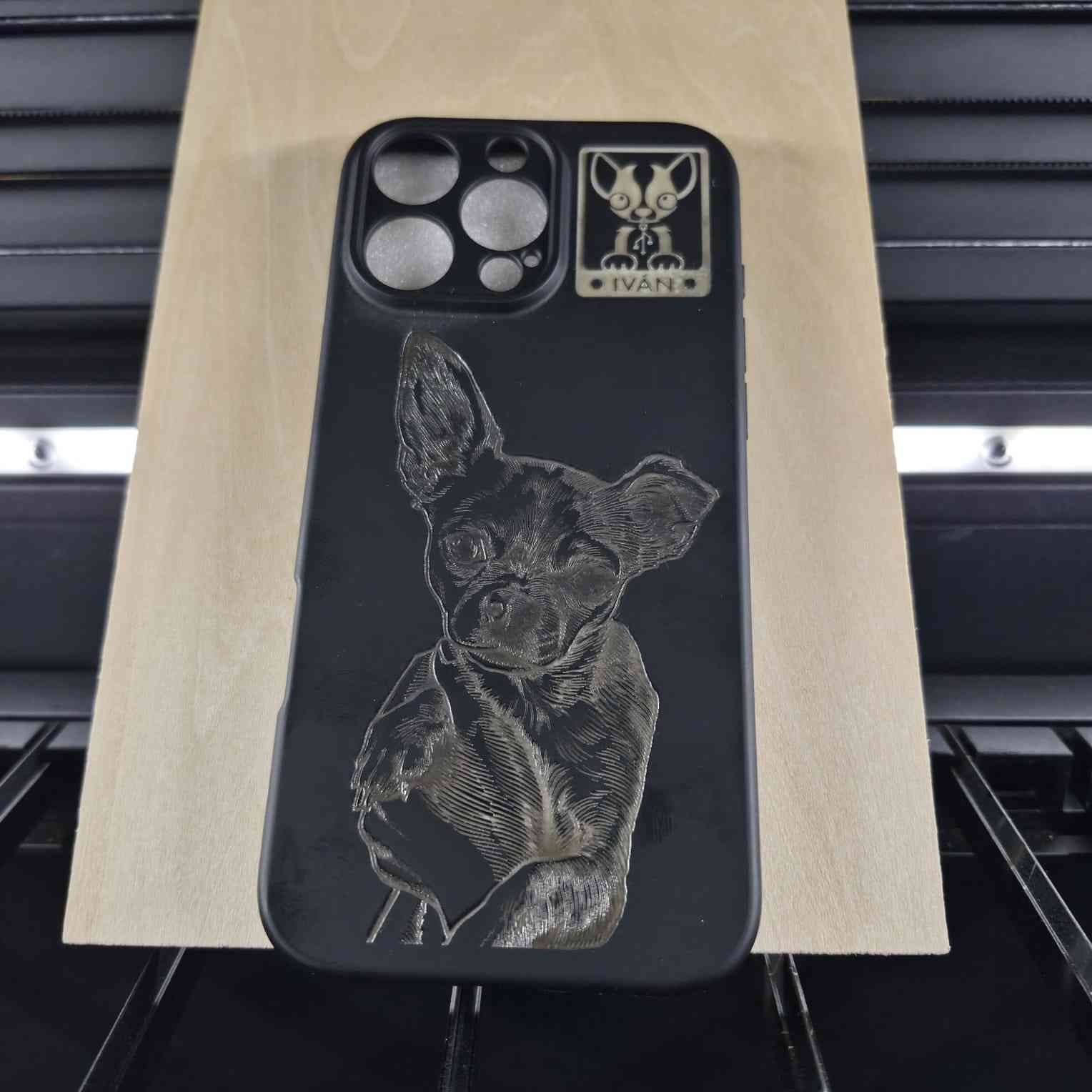 Funda de teléfono negra con grabado de perro apoyada sobre una superficie de madera dentro de una máquina de grabado láser.