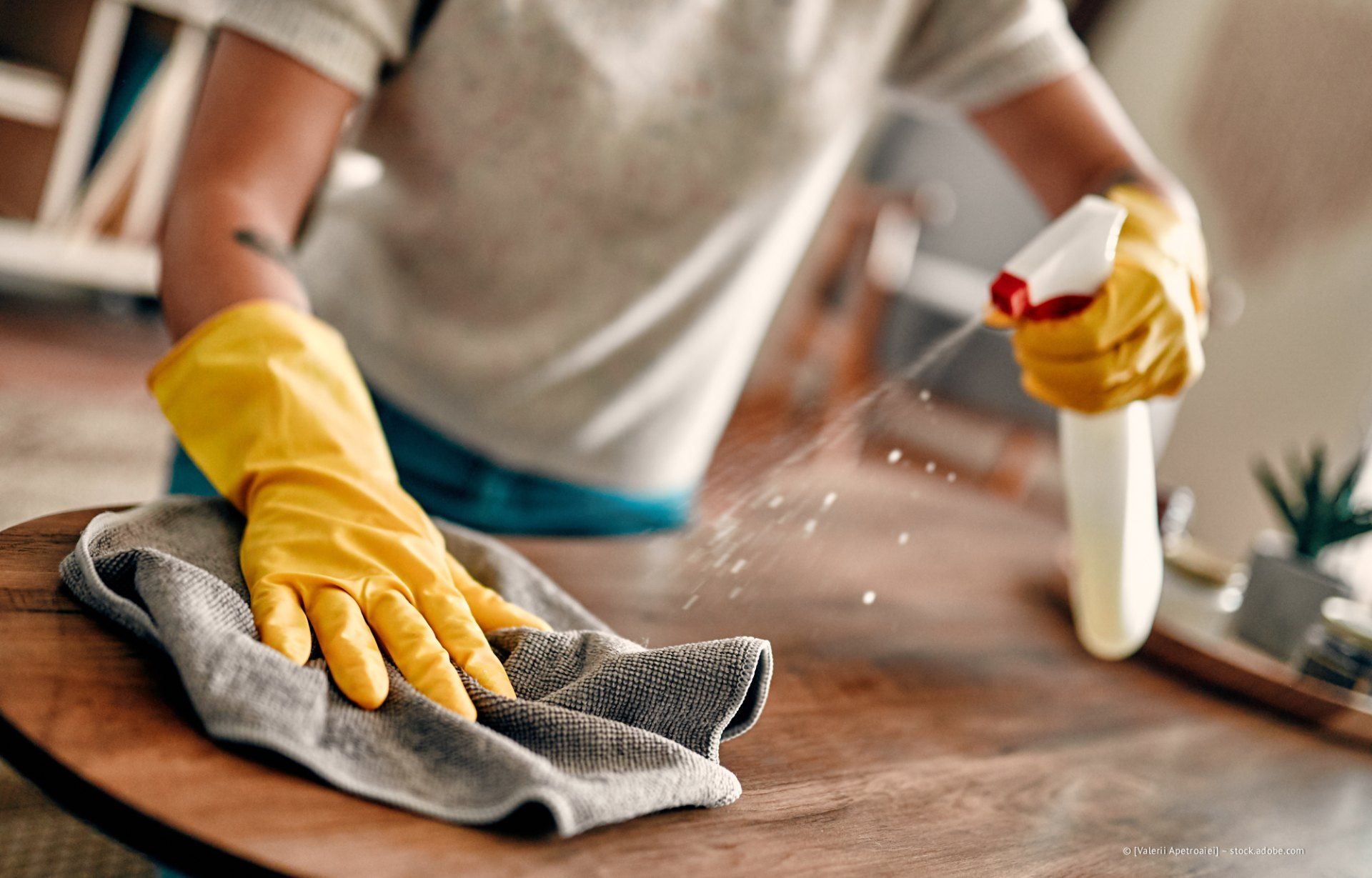 Eine Person, die gelbe Handschuhe trägt, sprüht Reinigungsmittel auf einen Holztisch und wischt ihn gleichzeitig mit einem grauen Tuch ab.