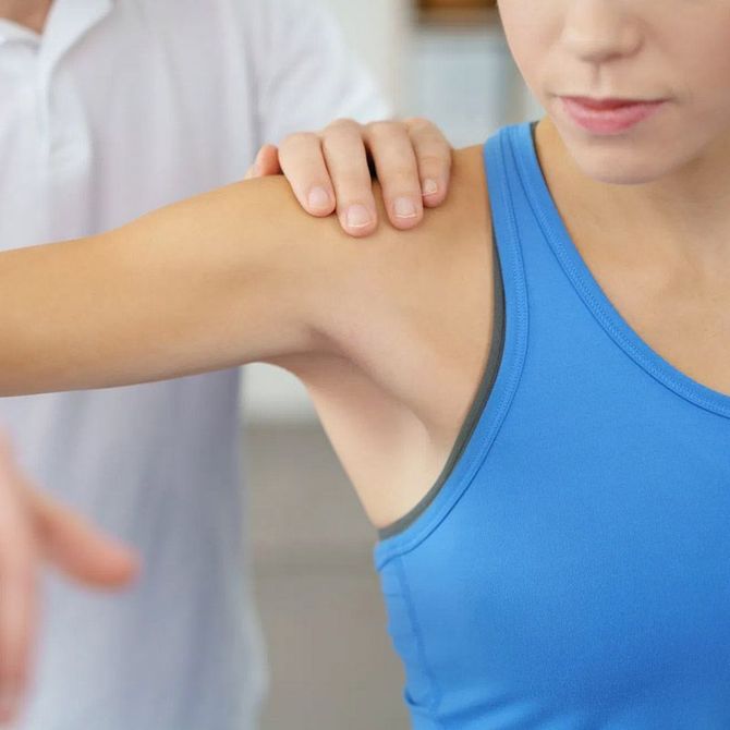 A una mujer que lleva una camiseta azul sin mangas un médico le examina el hombro.