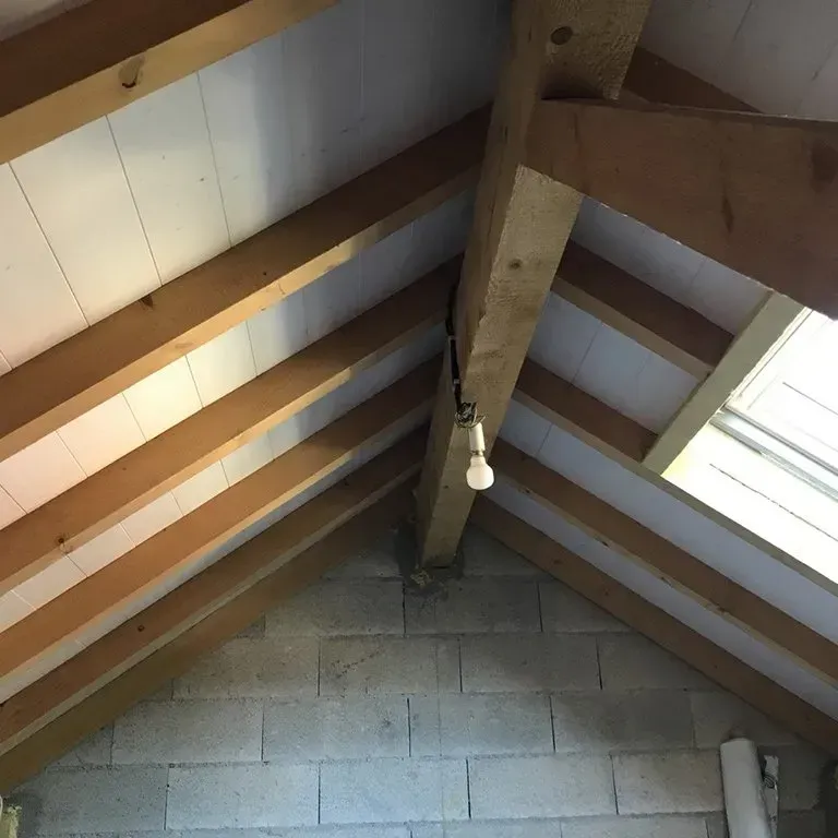 Intérieur de grenier avec des poutres apparentes en bois, un mur en parpaings et une ampoule suspendue.