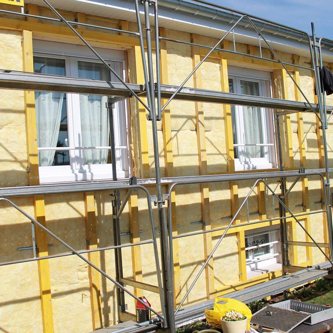 Façade d’une maison recouverte d’isolant jaune, avec un échafaudage et deux fenêtres équipées de rideaux, en cours de rénovation