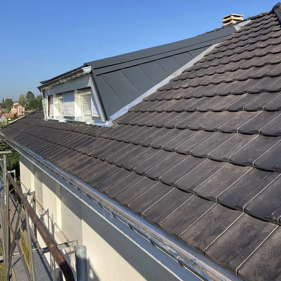Toit sombre en tuiles, avec des lucarnes et une extension en métal, sous un ciel bleu dégagé.