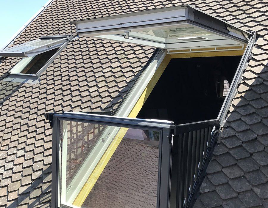 Fenêtre de toit ouverte sur une toiture en tuiles, avec une rambarde noire et un intérieur sombre visible