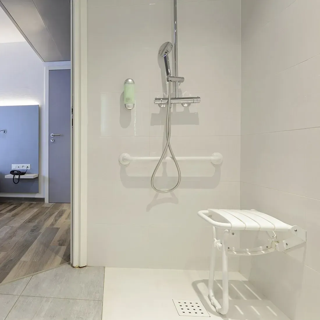 Douche accessible avec barres d’appui, douchette à main, siège rabattable et sol carrelé.