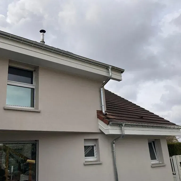 Maison beige à deux étages, avec des bordures blanches et un toit gris, sous un ciel nuageux