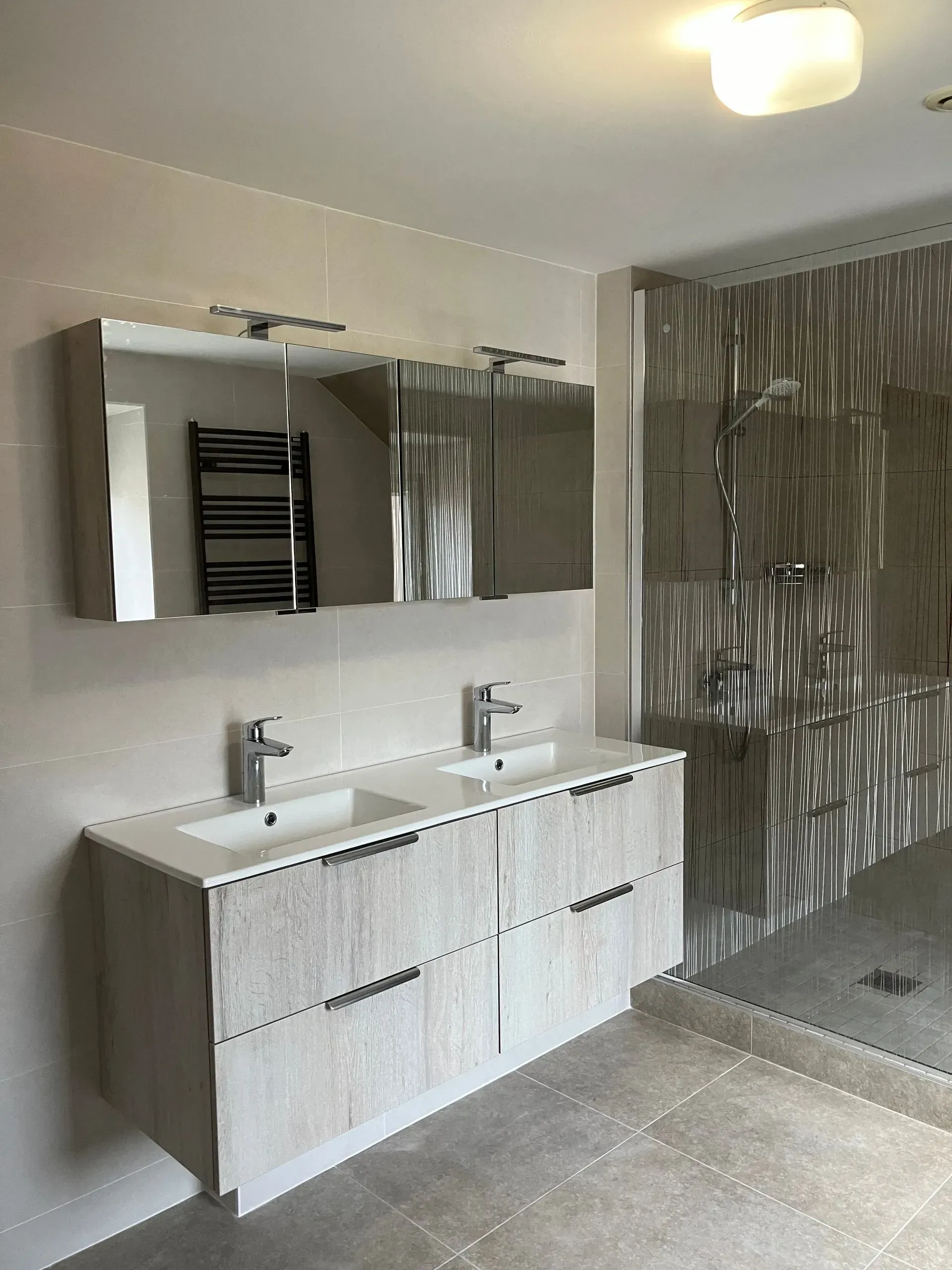 Salle de bains moderne avec double vasque, grand miroir et douche à l’italienne avec murs carrelés.