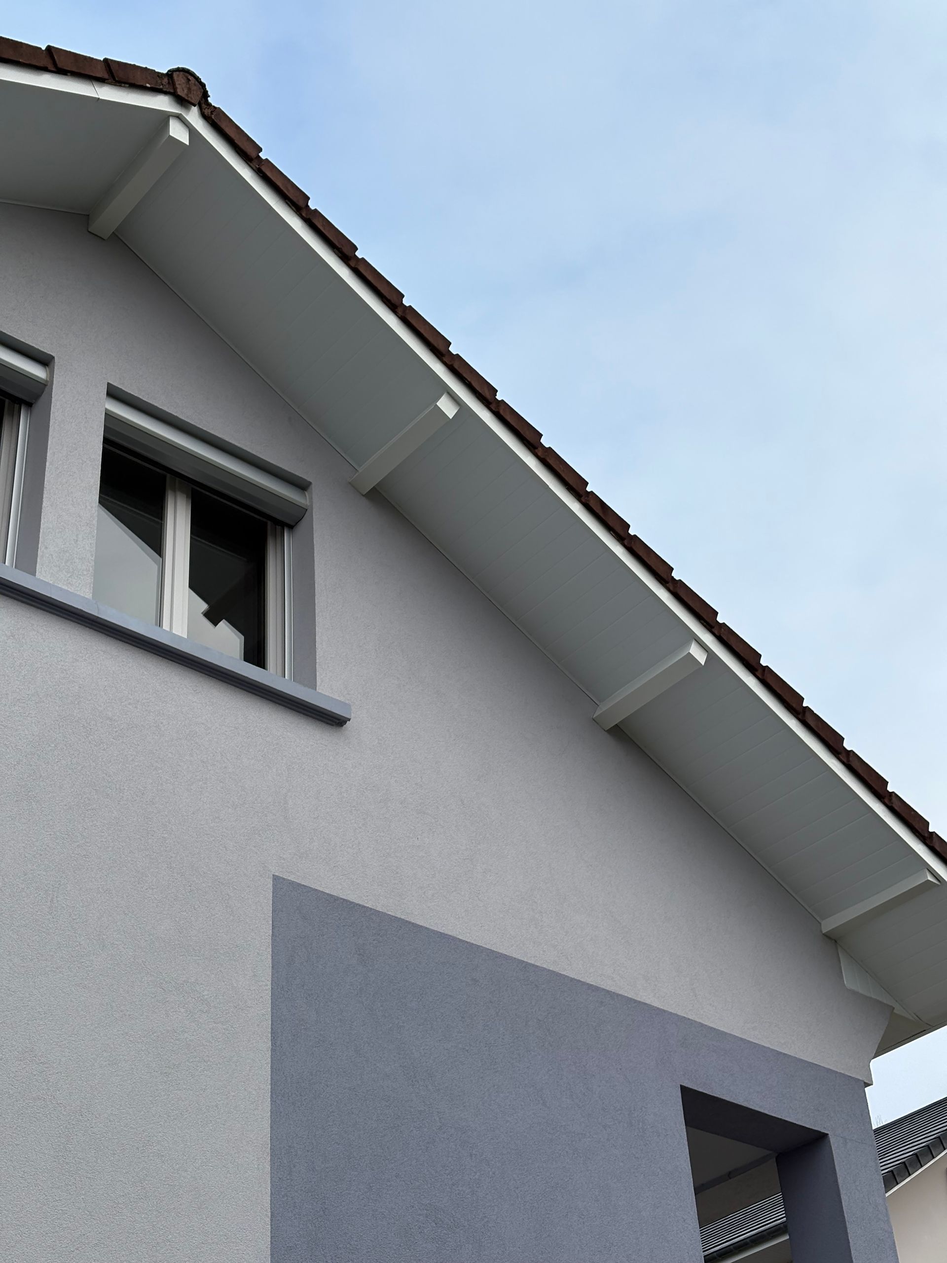 Extérieur d’une maison blanche avec un toit en pente, des murs gris et une fenêtre à l’étage, sur fond de ciel bleu