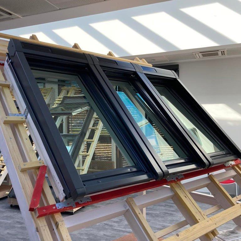 Grande fenêtre à cadre noir posée sur des tréteaux en bois dans un atelier lumineux, avec la lumière du soleil et des reflets.