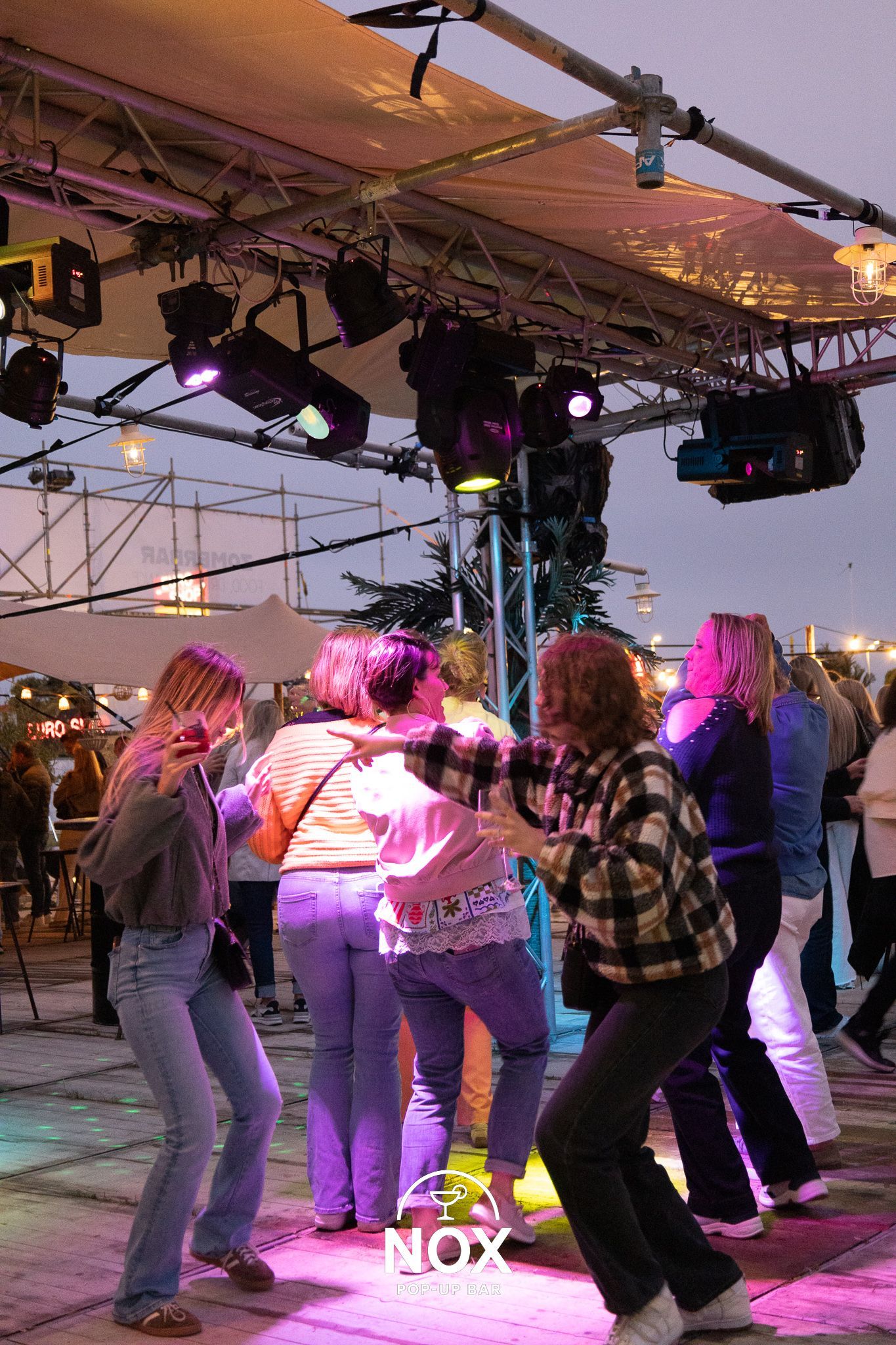 DJ in een volle nachtclub, met opgeheven armen en wijzend naar boven. Podium verlicht met blauwe en paarse lampen.