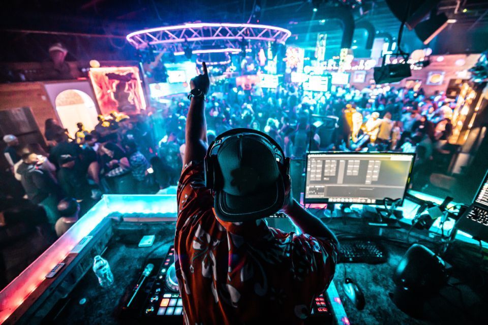 DJ in een volle nachtclub, met opgeheven armen en wijzend naar boven. Podium verlicht met blauwe en paarse lampen.