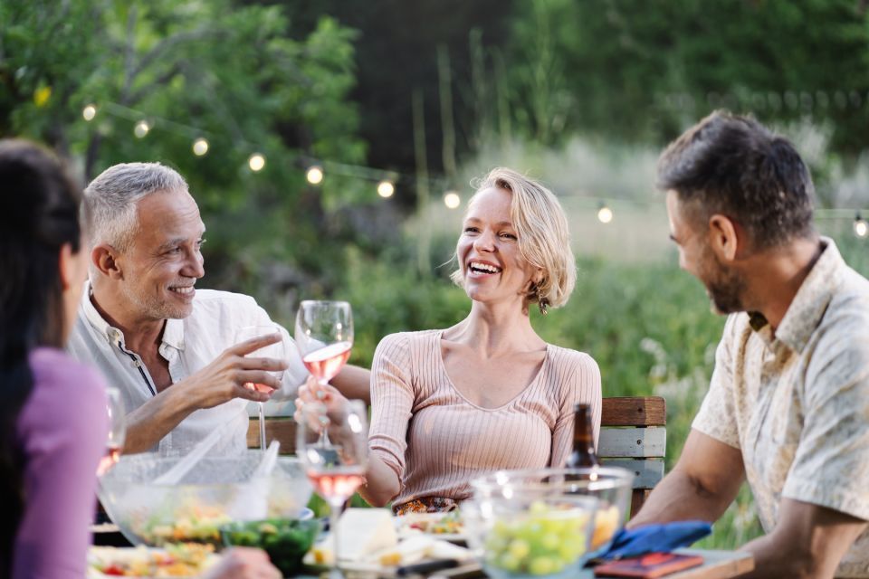 Mensen lachen en drinken wijn aan een eettafel in de buitenlucht, omringd door eten, lichtslingers en groen.