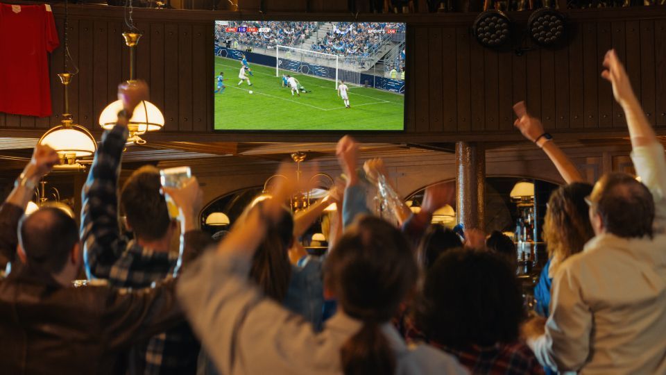 Mensen in een café juichen terwijl ze naar een voetbalwedstrijd op tv kijken. Ze hebben hun armen omhoog en houden drankjes vast.