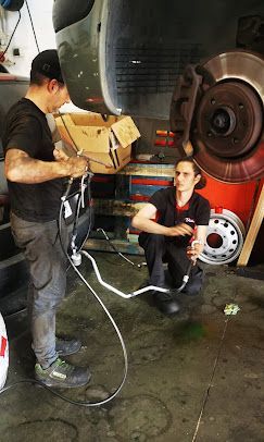 Dos mecánicos trabajando en un vehículo. Uno de pie, sujetando tubos; el otro, arrodillado, inspeccionando una manguera en un taller.