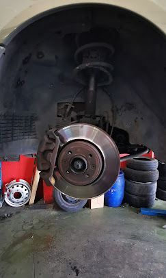 Disco y pinza de freno de automóvil, rueda quitada, mostrando la suspensión y la pila de neumáticos en un garaje.