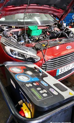 Un automóvil Ford rojo con el capó abierto, cerca de una máquina de servicio de aire acondicionado en un garaje.