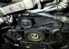 Primer plano de la correa serpentina y las poleas de un motor de automóvil, rodeadas de mangueras y otros componentes.