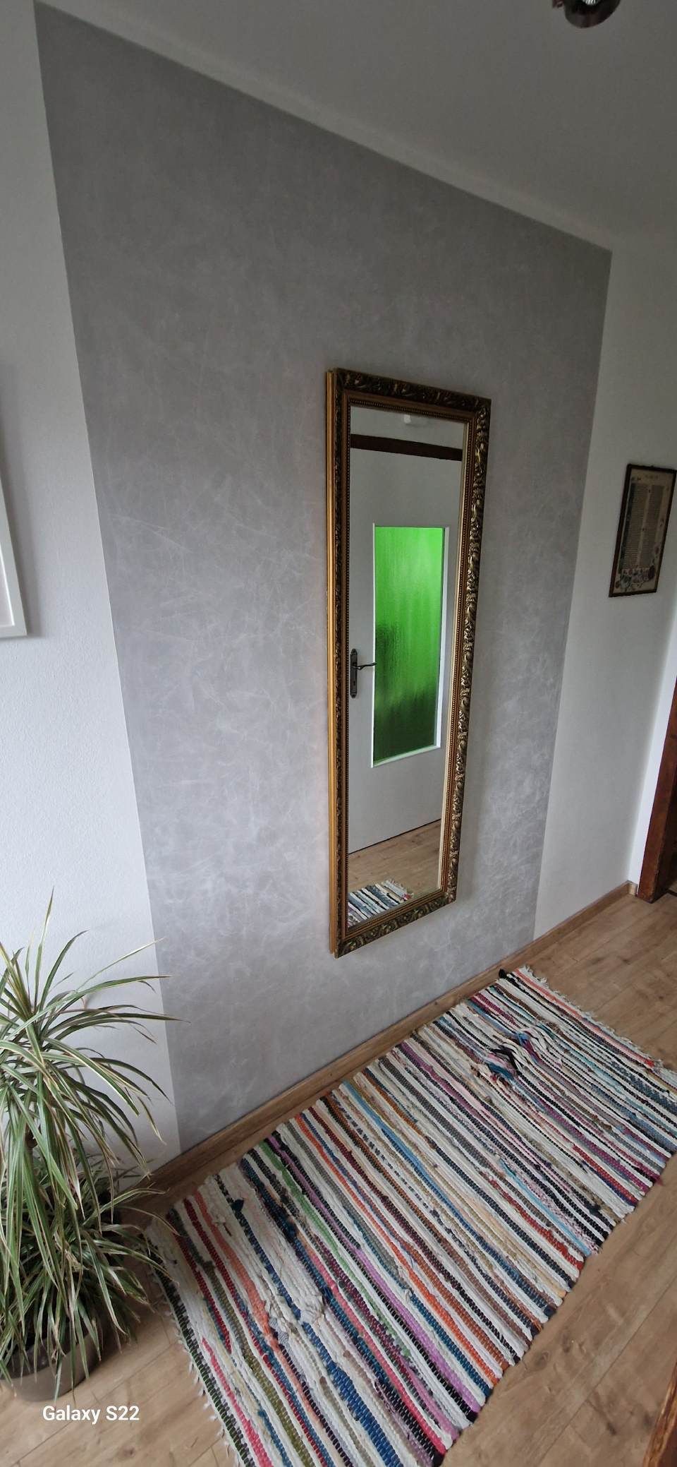 Flur mit einem farbenfrohen, gestreiften Läufer und einem an der Wand befestigten Spiegel, der ein grünes Türblatt reflektiert.