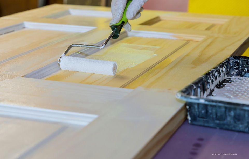 Eine behandschuhte Hand benutzt eine Farbrolle, um ein hölzernes Türblatt mit gelber Farbe zu streichen.