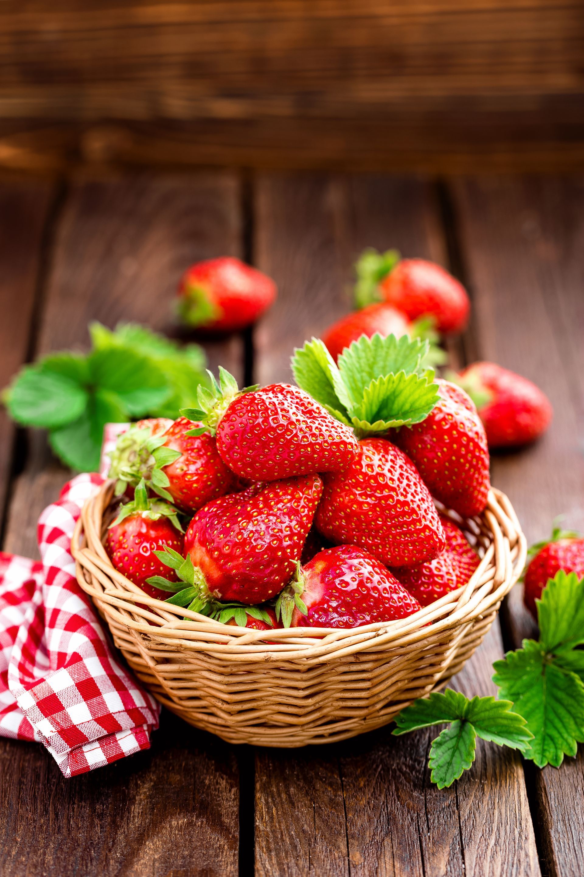 Un panier de fraises rouges mûres posé sur une table en bois, agrémenté de feuilles vertes fraîches et d'une serviette à carreaux.