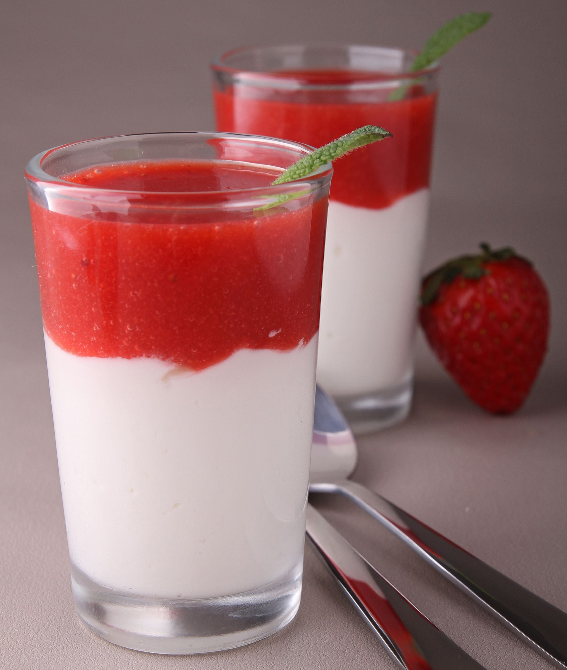 Deux verrines à dessert à étages ; base de crème blanche surmontée de purée de fraises rouges, garnie de menthe.