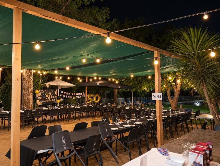 Terraza de restaurante al aire libre por la noche, con guirnaldas de luces, sillas negras y un toldo verde