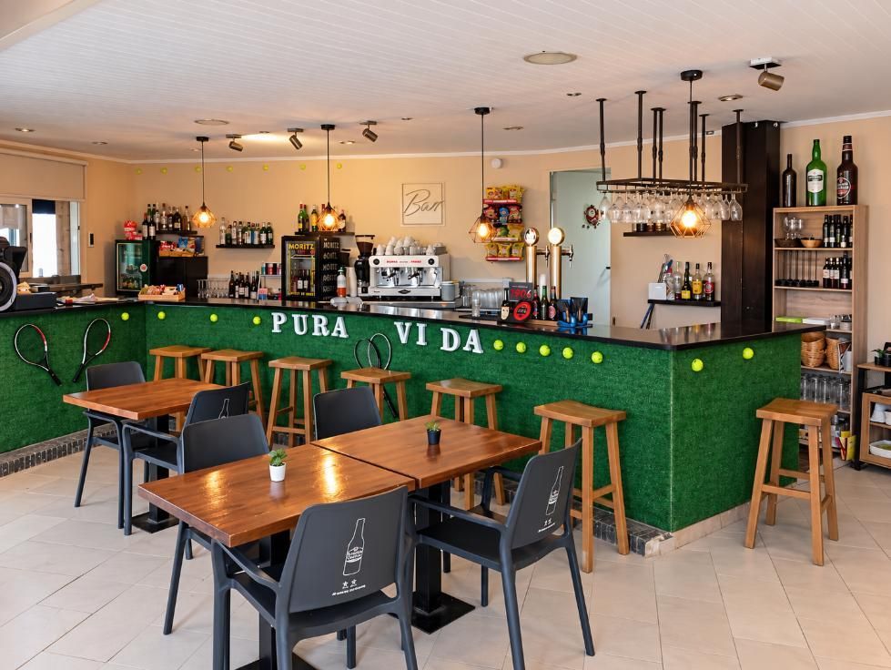 Interior luminoso de bar y cafetería, con barra verde, lámparas colgantes y mesas y taburetes vacíos