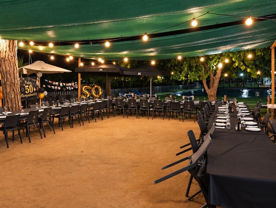 Zona de comedor al aire libre con guirnaldas de luces, mesas cubiertas de negro y sillas bajo una cubierta verde por la noche