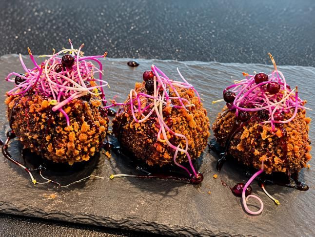 Tres croquetas crujientes de color marrón anaranjado, coronadas con brotes rosados, sobre una tabla de pizarra