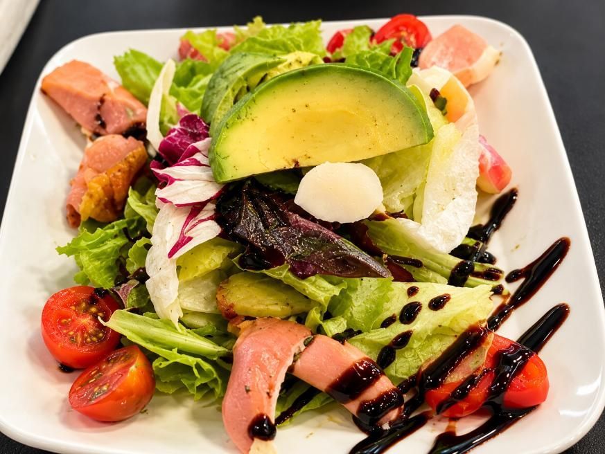 Ensalada con aguacate, hojas verdes, tomates, prosciutto y un toque de reducción de balsámico, servida en un plato blanco
