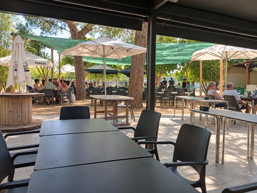 Terraza de cafetería al aire libre con mesas y sillas negras y sombrillas beige bajo una malla de sombreo verde