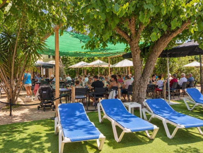 Zona de piscina sombreada de un resort, con tumbonas azules, árboles, sombrillas y gente sentada en mesas al aire libre