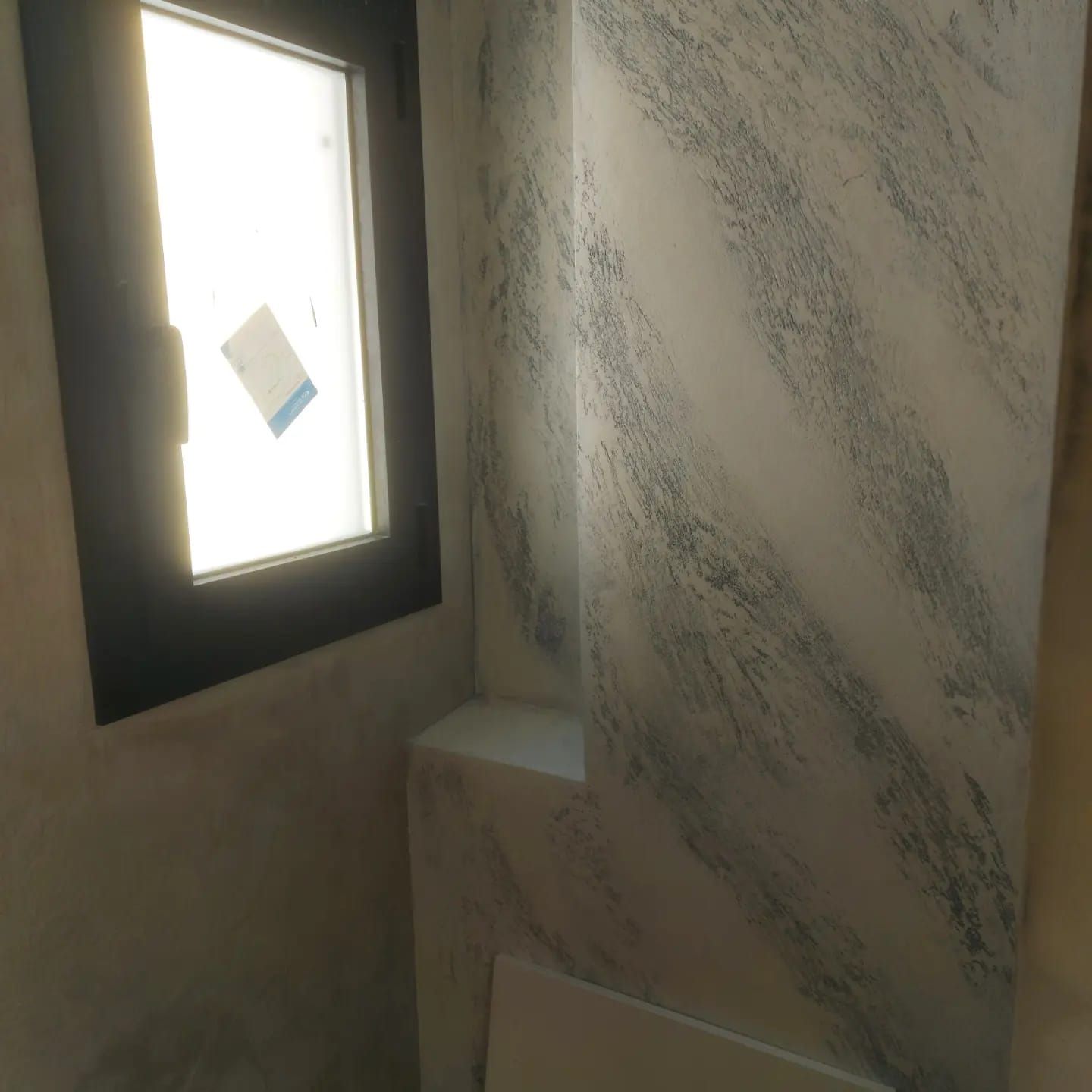Ventana pequeña en una pared con textura, encima de un lavabo blanco en una habitación alicatada