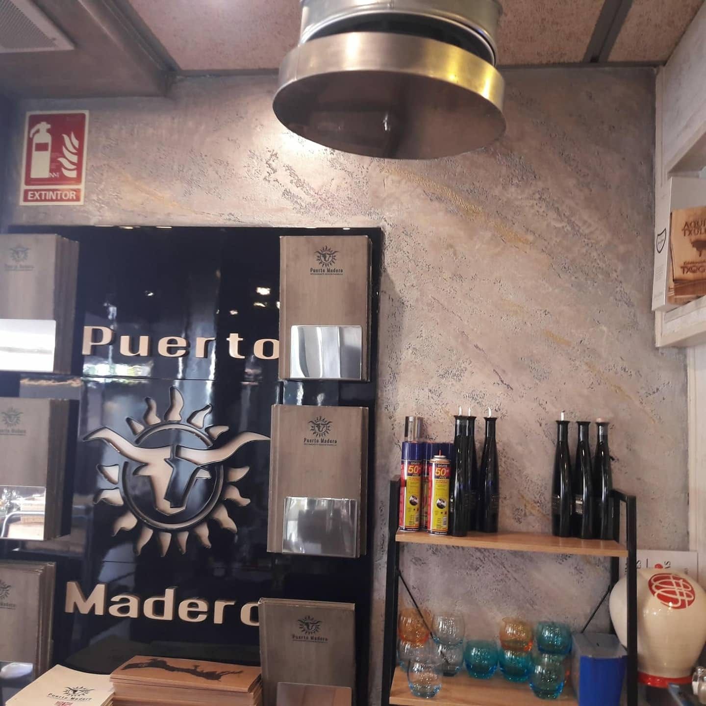 Pared de café rústico con un letrero de Puerto Madero, dispensadores metálicos y estantes con botellas y cristalería
