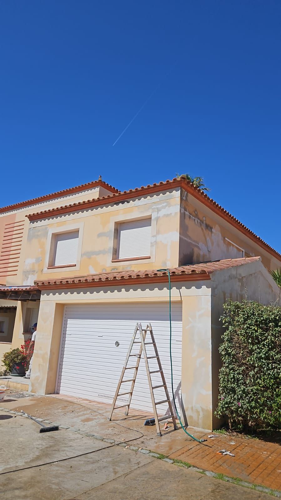 Casa de estuco de dos plantas con garaje, una escalera apoyada en la puerta y un cielo azul despejado.