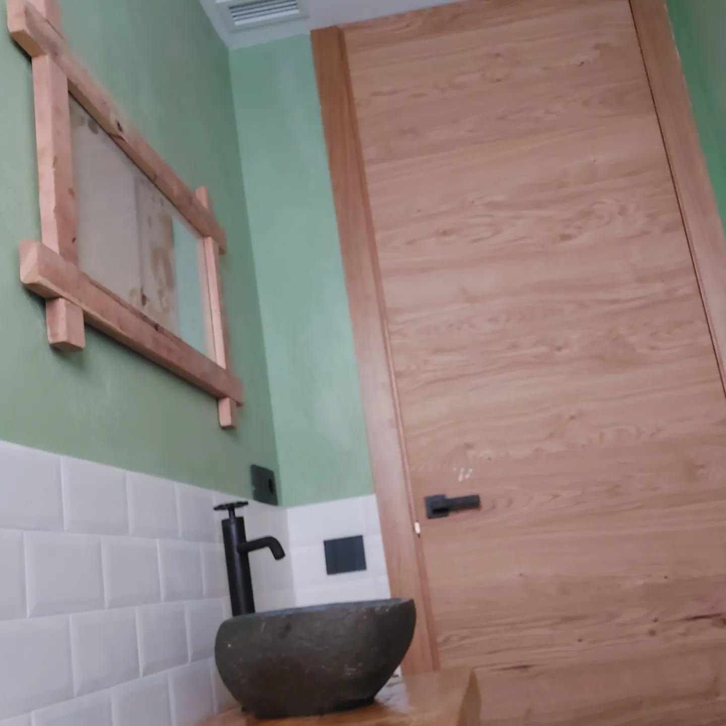 Baño pequeño con paredes verdes, azulejos blancos, marco de espejo de madera y un lavabo sobre encimera.