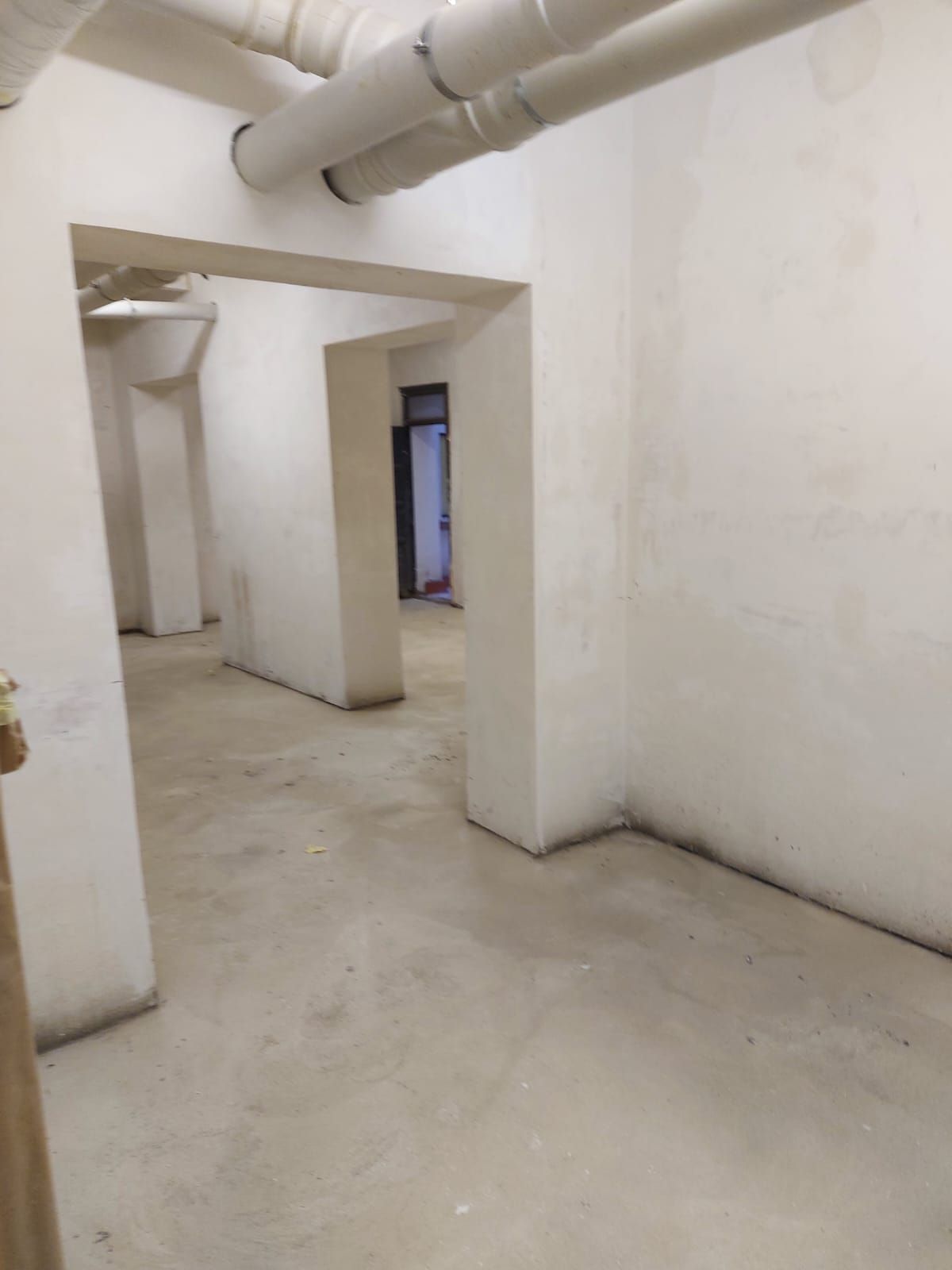 Pasillo estrecho de hormigón blanco, con tuberías vistas en el techo y puertas que dan a una habitación iluminada.